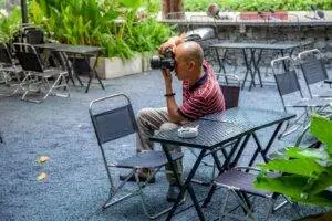 Man photographing at an outdoor cafe surrounded by empty chairs.