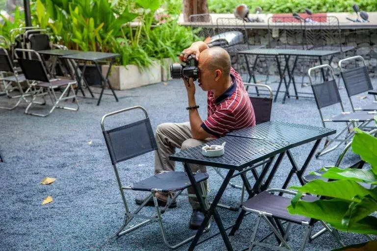 Man photographing at an outdoor cafe surrounded by empty chairs.